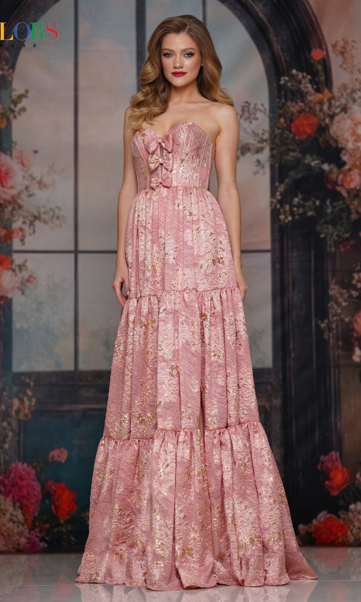 A woman stands before a floral and window backdrop, wearing the Colors Dress 3794—a strapless long prom dress featuring a corset bodice and a pink, floral-patterned tiered skirt with lace details.